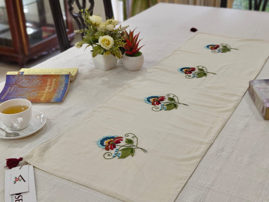 Embroidered table runner with floral designs on a dining table with books and a cup.