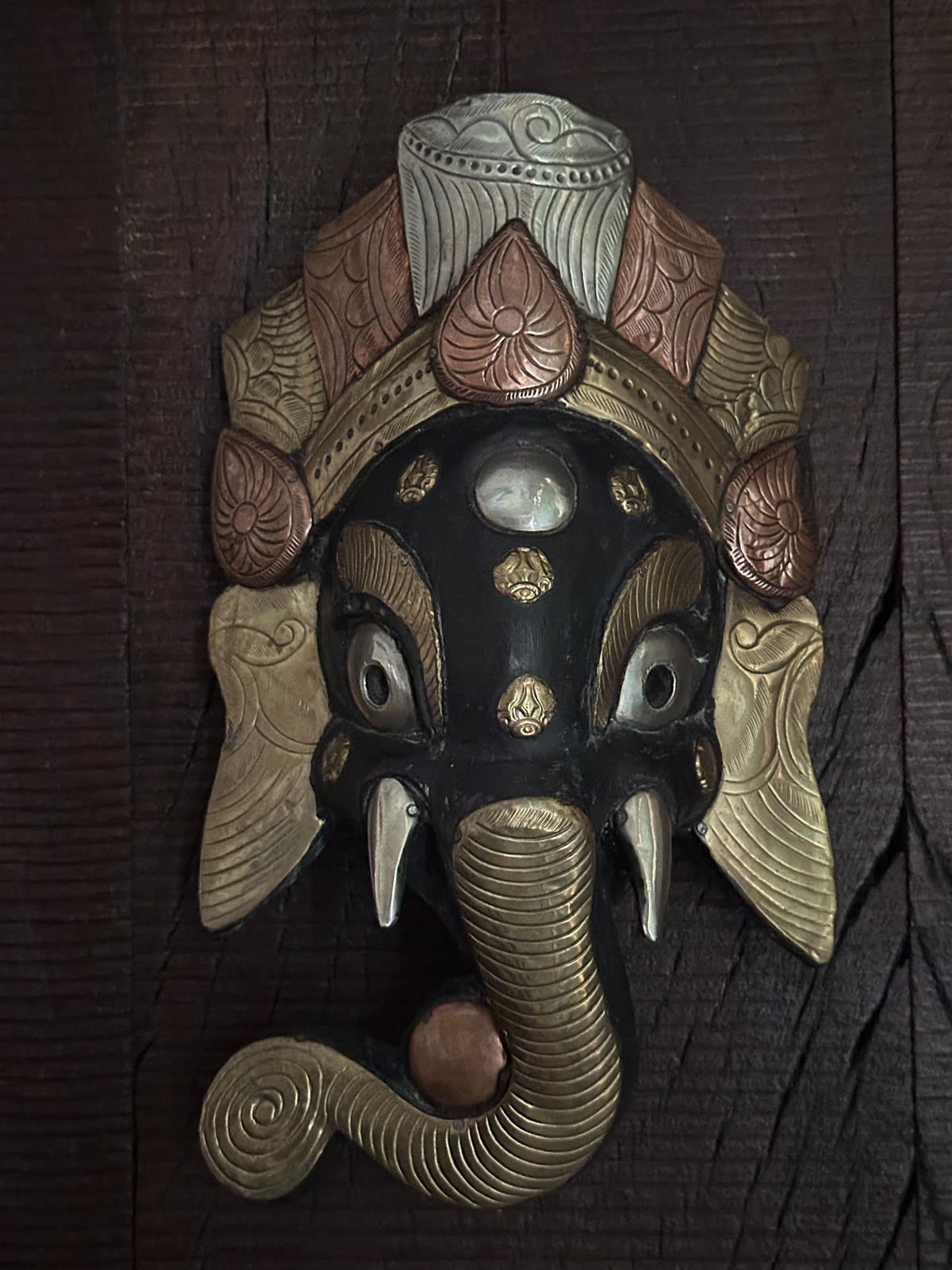 A decorative Ganesha wall hanging or mask with a black face, copper, brass and metal-plated ears, trunk, eye areas, and headdress details, mounted against a dark wood background.